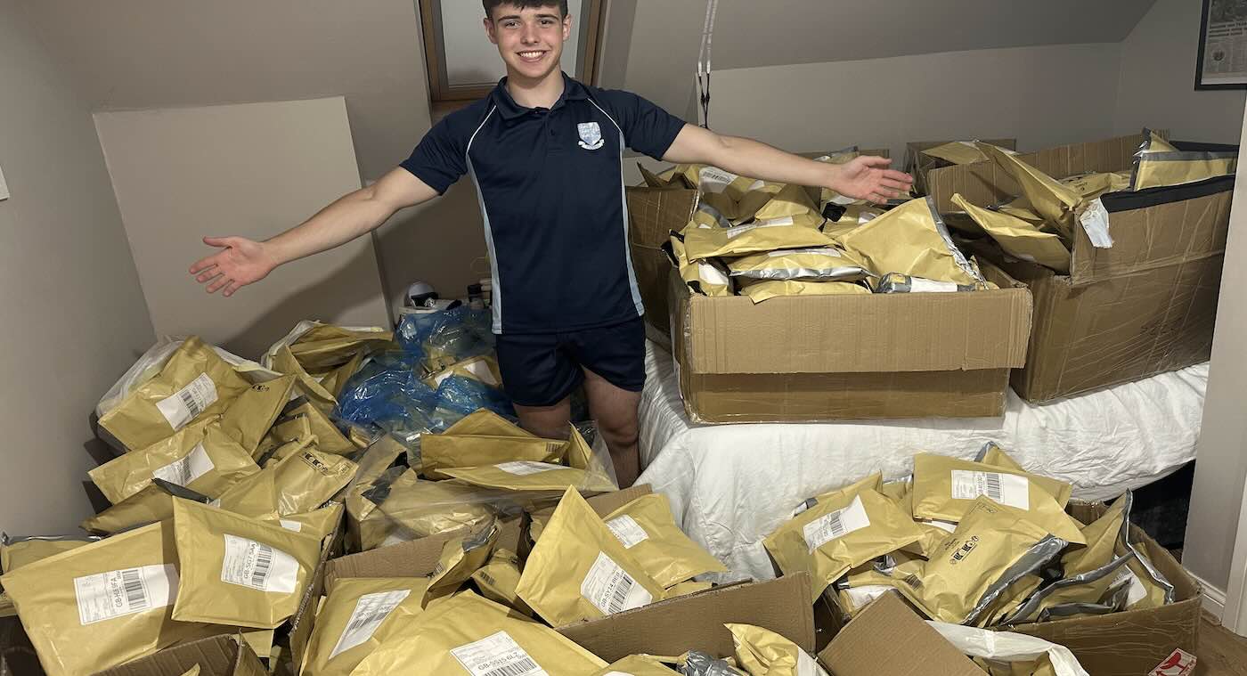 Harrison Nott stands on his bed surrounded by hundreds of gold courier packages ready to ship, arms spread wide, grinning