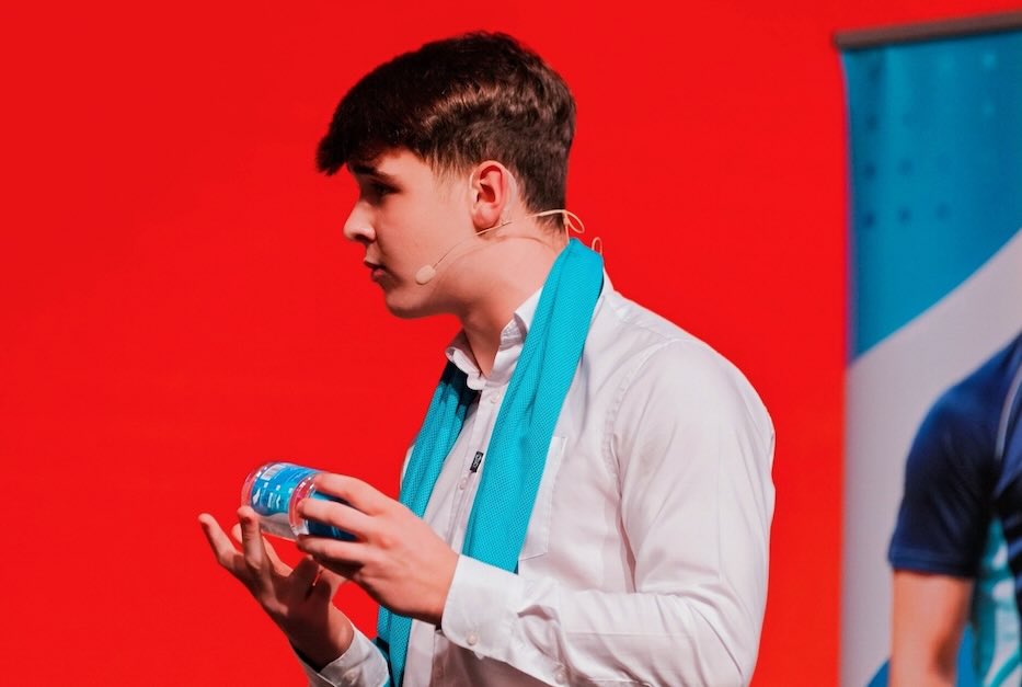Harrison Nott on stage at Alibaba CoCreate Pitch 2025 London, CoolTowel draped around his neck, holding the product to the audience, red backdrop